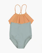 Der Ava Mädchen Badeanzug von Main Design ist ein Kinderbadeanzug mit einem hellblauen Unterteil und einem pfirsichfarbenen Rüschenoberteil. Er hat dünne, gekreuzte Träger auf der Rückseite, was ihn schlicht und zum Schwimmen geeignet macht. Der Hintergrund ist schlicht weiß.