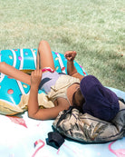 Das Kind trägt den "Ava Mädchen Badeanzug" von Main Design in Lila, entspannt auf einem bunten Pool-Float im Gras, Snack in der Hand. Daneben: Palm-Print-Tasche und Flamingo-Handtuch. Der Badeanzug hat verstellbare Träger.