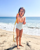 Ein kleines Kind auf dem Ava Mädchen Badeanzug von Main Design steht barfuß an einem Sandstrand und hält ein langes Stück Seetang hoch, mit dem Meer und dem blauen Himmel im Hintergrund.