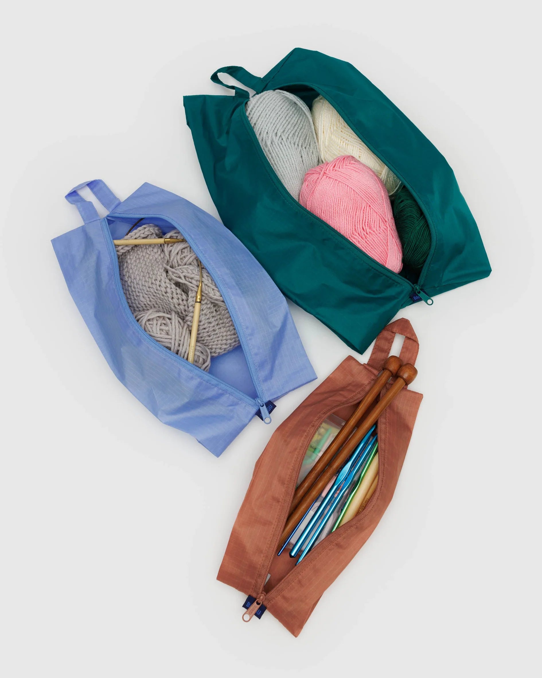 Das BAGGU 3D Zip Set von BAGGU besteht aus blauen, grünen und braunen Reißverschlusstaschen für Strickutensilien auf einer weißen Oberfläche - ideal als umweltfreundliche Taschen oder praktische Reisebegleiter.