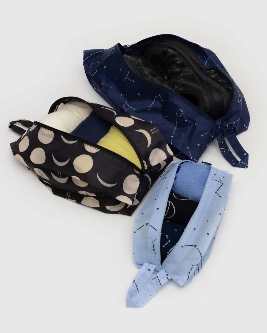 Abgebildet sind drei gemusterte BAGGU 3D Zip Set Organizer von BAGGU: ein dunkelblauer mit Sternbildern für Schuhe, ein schwarzer mit Mondphasen für Kleidung und ein hellblauer mit Sternen bedruckter aufklappbarer Organizer - perfekt für organisiertes, stilvolles Reisen.