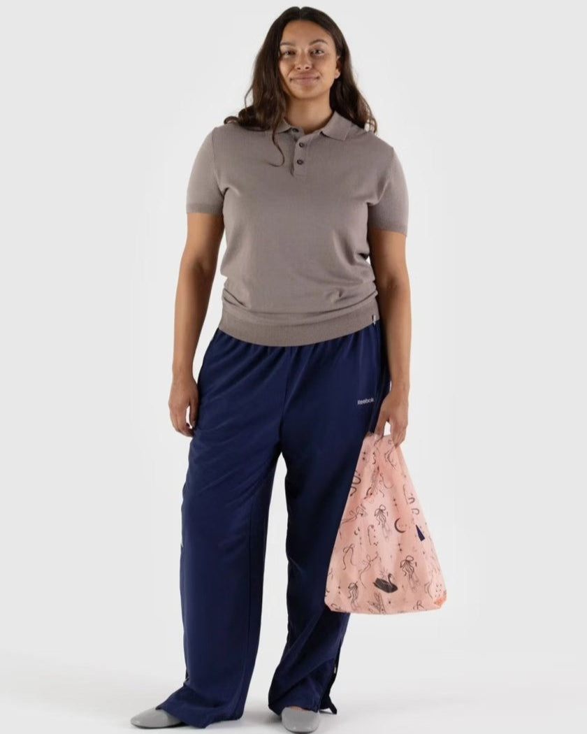 Eine Frau steht vor einem schlichten Hintergrund, trägt ein taupefarbenes kurzärmeliges Polo, eine weite marineblaue Hose, graue Schuhe und hält eine BAGGU Baby Tragetasche aus recyceltem Nylon mit abstrakten Linienzeichnungen in der Hand.