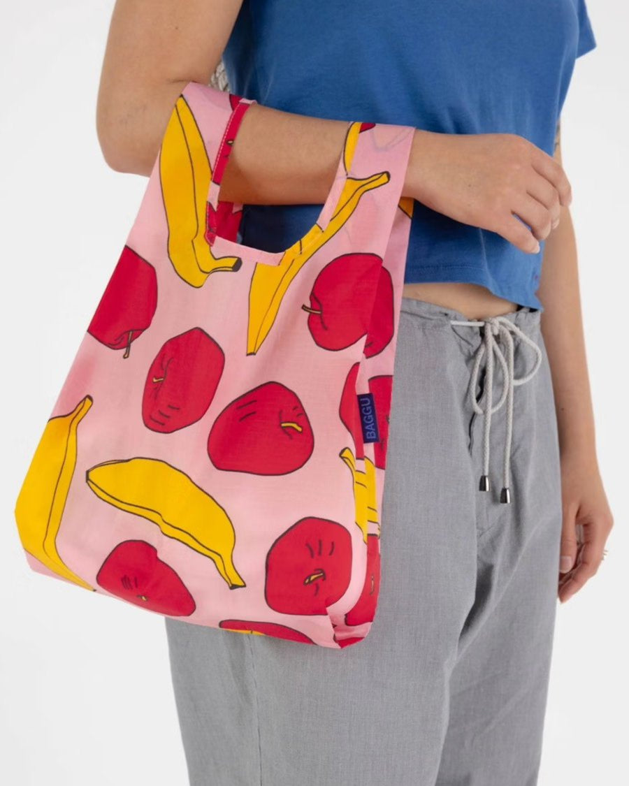 Eine Person mit blauem Top und grauer Hose hält die umweltfreundliche BAGGU Baby Tragetasche aus recyceltem Nylon mit bunten Apfel- und Bananenmotiven, kurzen Henkeln und farbenfrohem Design von BAGGU.