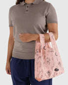 Eine Person in einem taupefarbenen kurzärmeligen Polo und einer marineblauen Hose hält eine BAGGU Baby Tragetasche, blassrosa mit schwarzen Abbildungen von Schlangen, Quallen, Monden, UFOs und Sternen.