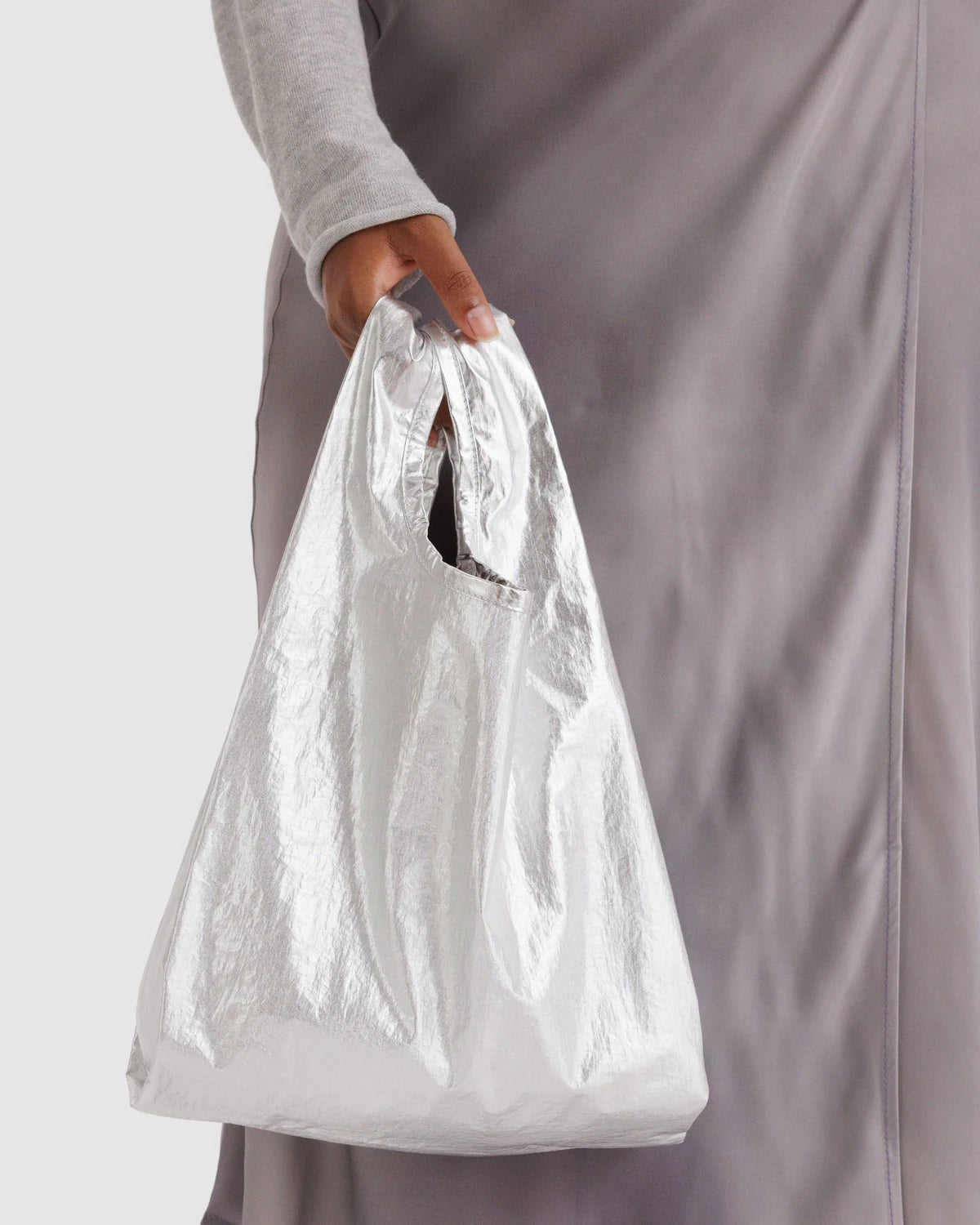 Eine Person in einem hellgrauen, langärmeligen Oberteil und einem seidengrauen Rock hält die BAGGU Baby Tragetasche, eine silbern glänzende Tasche aus recyceltem Nylon von BAGGU mit einem umweltfreundlichen, ausgeschnittenen Griffdesign.