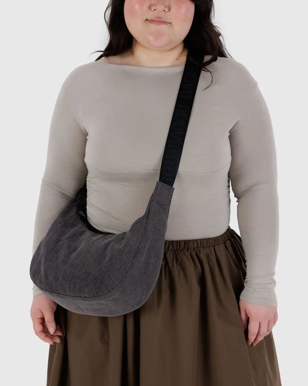 Eine Person in einem beigen Oberteil und einem braunen Rock trägt die BAGGU Medium Denim Crescent Bag mit einem schwarzen Riemen über der Brust. Der schlichte weiße Hintergrund unterstreicht dieses nachhaltige BAGGU-Accessoire.