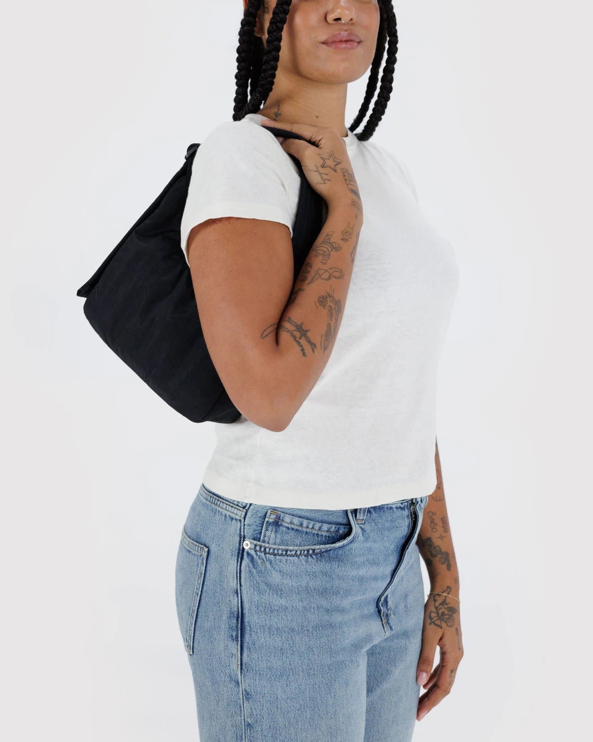 Eine Person mit geflochtenem Haar und Tattoos trägt ein weißes T-Shirt und blaue Jeans und hält die BAGGU Mini Nylon Shoulder Bag. Der schlichte weiße Hintergrund unterstreicht dieses stilvolle, umweltfreundliche Accessoire von BAGGU.