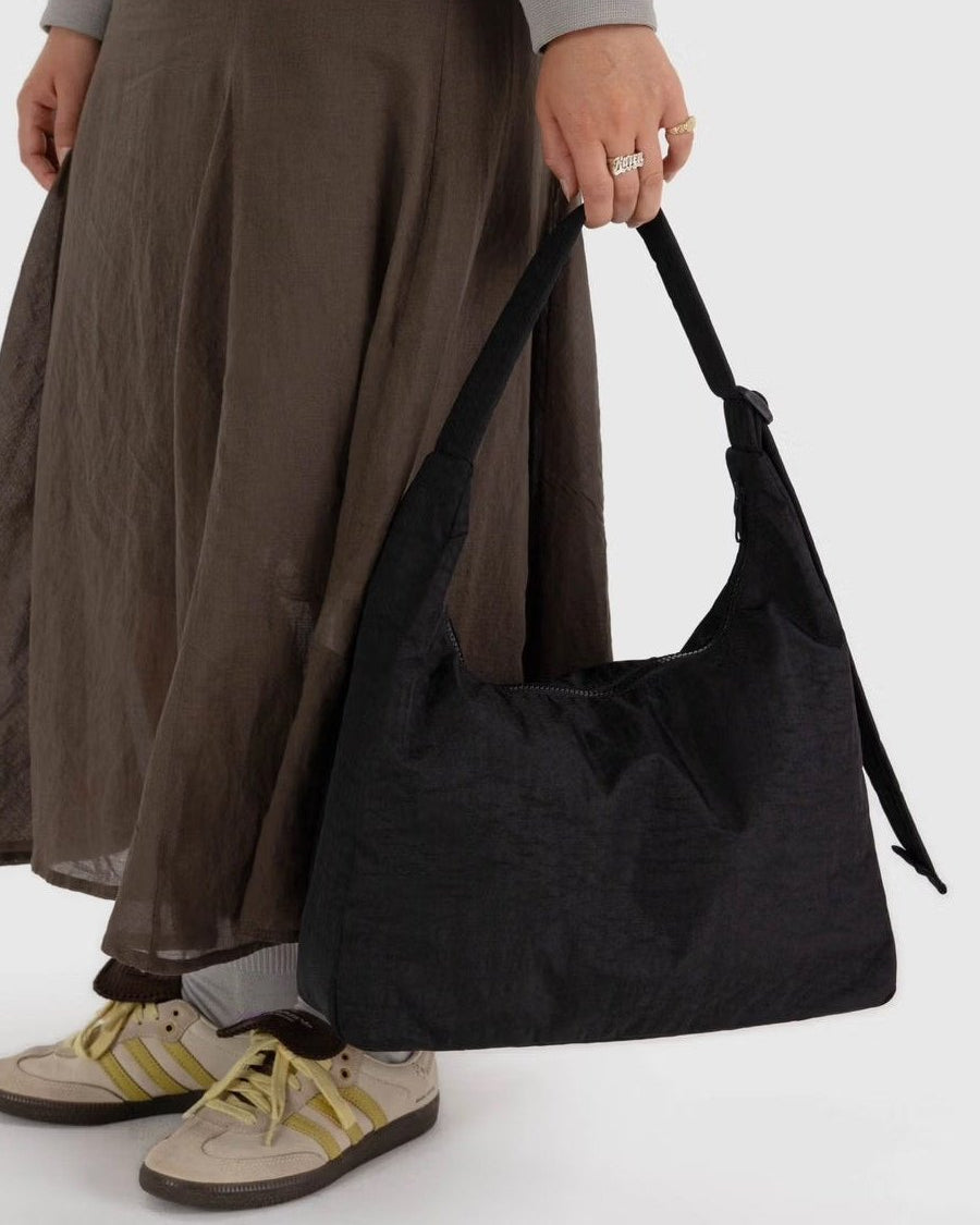 Eine Person in einem fließenden braunen Rock und cremefarbenen Turnschuhen mit gelben Streifen hält eine große BAGGU-Nylon-Umhängetasche, ihre Hand mit mehreren Ringen ruht auf dem Griff.