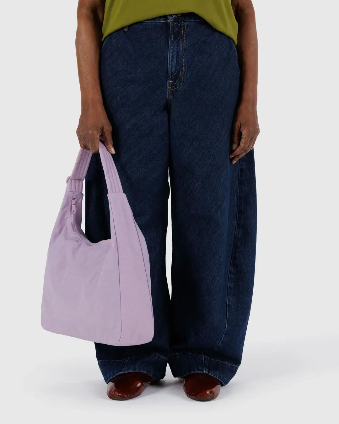 Eine Person steht vor einem hellen Hintergrund und zeigt nur ihren Unterkörper in einer blauen Jeans mit weitem Bein, einem grünen Oberteil und braunen Schuhen, während sie eine pastellviolette BAGGU-Nylon-Schultertasche von BAGGU hält.