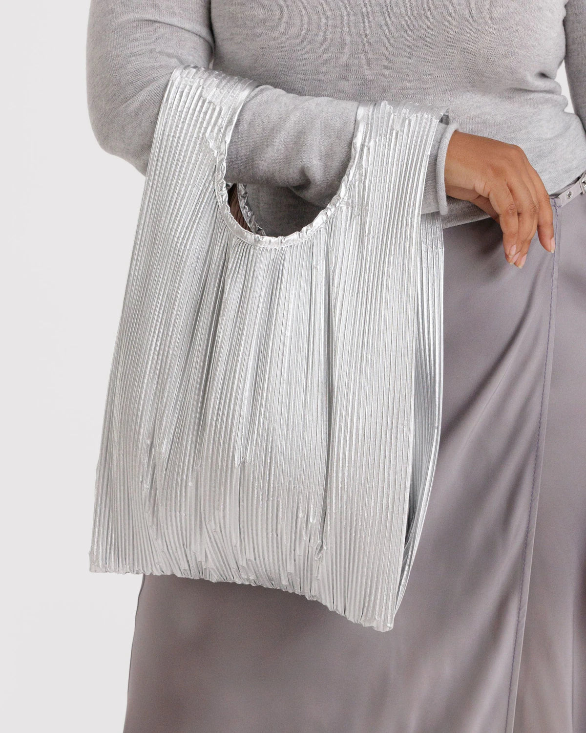 Eine Person in einem hellgrauen, langärmeligen Oberteil und einem seidengrauen Rock hält die BAGGU Pleated Tragetasche von BAGGU, eine metallisch-silberne Allround-Tasche mit runden Henkeln, die aus recyceltem Ripstop-Nylon für umweltfreundlichen Stil hergestellt ist.