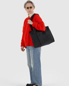 Ein älterer Erwachsener mit kurzen grauen Haaren, Sonnenbrille, rotem Pullover, hellblauen Jeans und schwarzen Schuhen steht mit einer BAGGU Small Cloud Carry-On Tasche von BAGGU über der Schulter vor einem einfachen hellen Hintergrund.