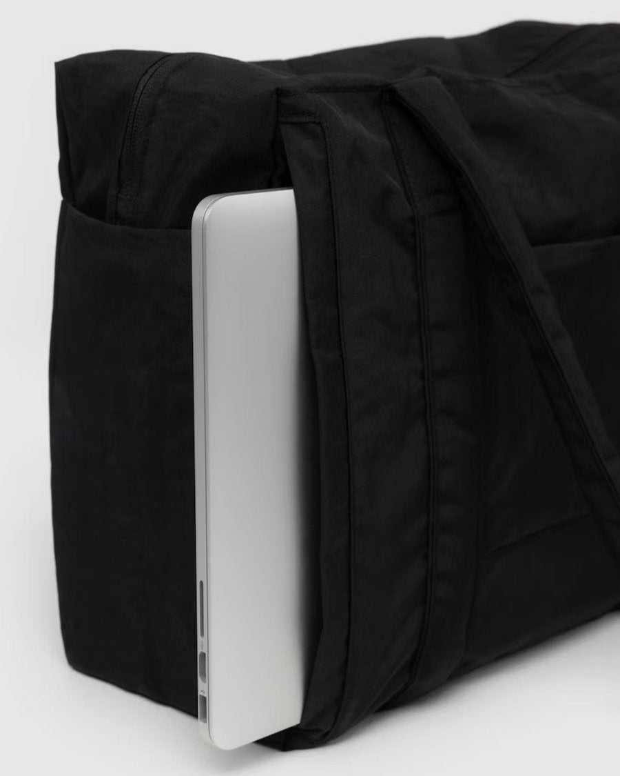 Ein silberner Laptop passt teilweise in die Seitentasche einer schwarzen BAGGU Small Cloud Carry-On Tasche, deren Schultergurte vor einem schlichten hellen Hintergrund zu sehen sind.