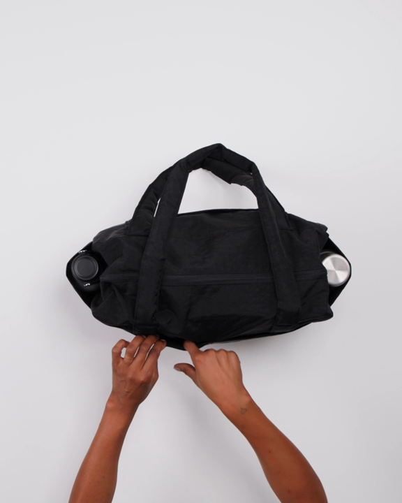 Eine Person öffnet den Reißverschluss einer BAGGU Small Cloud Carry-On Tasche aus recyceltem Nylon, die auf einer weißen Fläche steht. Die BAGGU Tasche hat zwei Griffe und Metallwasserflaschen in den Seitentaschen.