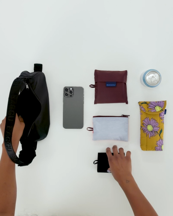 Eine Person legt eine BAGGU Small Denim Crescent Bag, ein Smartphone, eine Dose, drei kleine Taschen und eine gelbe Blumentasche auf eine weiße Fläche. Eine Hand greift nach einem schwarzen Beutel, während die andere die BAGGU-Tasche hält.