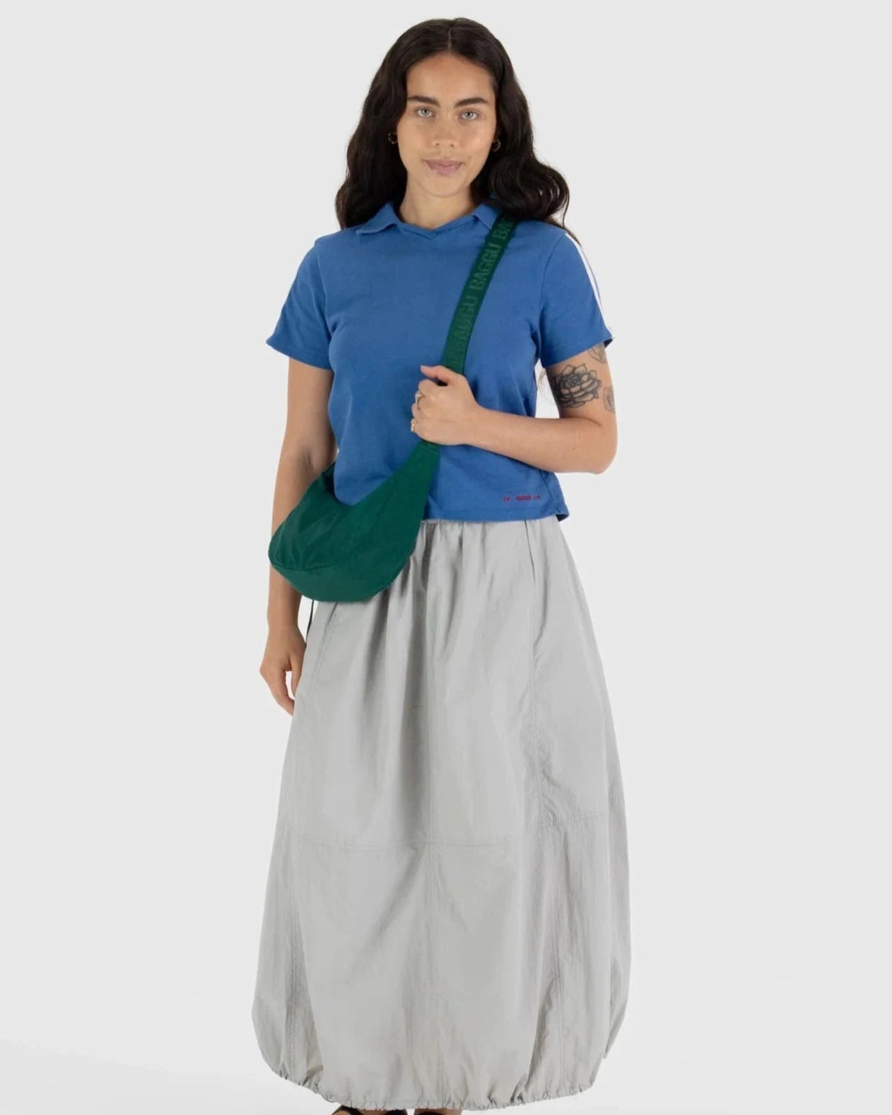 Eine Frau mit langen dunklen Haaren trägt ein blaues Poloshirt, einen hellgrauen, knöchellangen Rock und trägt die BAGGU Small Nylon Crescent Bag. Sie steht mit dem Gesicht zur Kamera vor einem schlichten weißen Hintergrund.