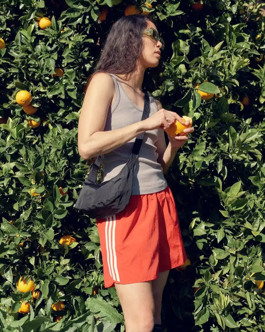 Eine Frau mit Sonnenbrille, einem grauen Tanktop, roten Shorts und einer schwarzen BAGGU Small Nylon Crescent Bag steht vor einem Orangenbaum und schält und isst eine Orange, während das Sonnenlicht sie und die grünen Blätter beleuchtet.