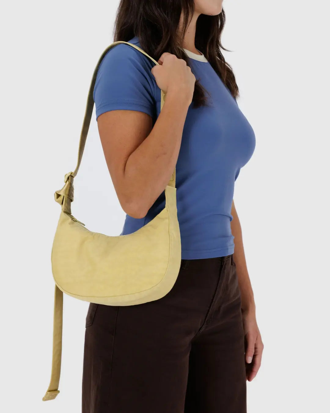 Eine Frau in einem blauen T-Shirt und einer dunkelbraunen Hose trägt die BAGGU Small Nylon Crescent Bag über der Schulter. Der schlichte, helle Hintergrund unterstreicht die umweltfreundliche Ausstrahlung dieses BAGGU-Accessoires.