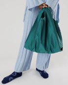 Eine Person in hellblauem, geripptem Pyjama und dunkelblauen Socken mit weißem Muster hält eine grüne BAGGU Standard Tragetasche von Main Design aus recyceltem Ripstop-Nylon. Der Hintergrund ist schlicht und hell.