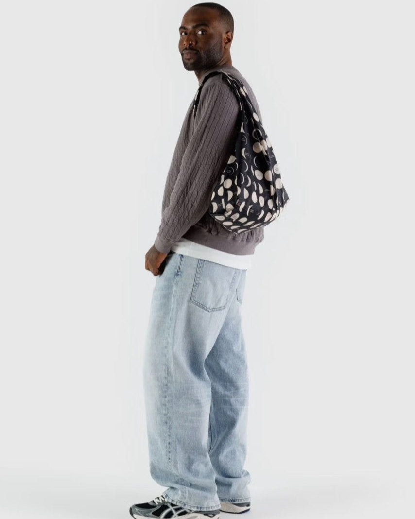 Ein Mann in grauem Strukturpullover, weißem T-Shirt, hellblauen Baggy-Jeans und grauen Turnschuhen steht seitlich mit einer BAGGU Standard Tragetasche von BAGGU aus recyceltem Ripstop-Nylon vor einem schlichten hellen Hintergrund.