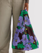 Eine Person in hellgrauer Hose hält die BAGGU Standard Tragetasche von BAGGU, eine nachhaltige Tasche aus recyceltem Ripstop-Nylon mit einem lebhaften lila und grünen Blumenmuster vor einem schlichten weißen Hintergrund.