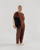 Ein älterer Mann mit kurzen grauen Haaren lächelt vor einem schlichten Hintergrund. Er trägt ein braunes T-Shirt, ein beiges langärmeliges Hemd, eine passende Hose, cremefarbene Socken und hat die BAGGU Zip Duck Bag von BAGGU aus recycelter Baumwolle dabei.