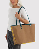 Eine ältere Frau mit langen grauen Haaren, gekleidet in ein weißes Oberteil und eine graue Hose, trägt die BAGGU Large Nylon Reversible Tote – mit blaugrünen Henkeln und gelber Einfassung – über der Schulter, während sie vor einem hellen Hintergrund steht.
