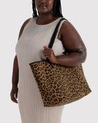 Eine Frau in einem ärmellosen beigen Kleid trägt die BAGGU Large Nylon Reversible Tote mit auffälligem Leopardenmuster vor einem schlichten hellen Hintergrund über der Schulter.