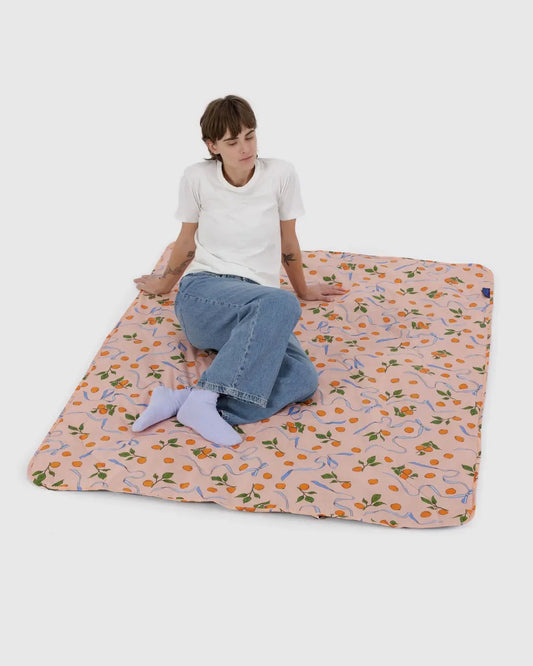 Auf einer großen rosafarbenen BAGGU Puffy Picknickdecke von Main Design, die mit Blumen- und Fruchtmustern verziert ist, sitzt eine Person in weißem T-Shirt und Blue Jeans mit gekreuzten Beinen und lila Socken zurückgelehnt. Diese nachhaltige Decke aus recyceltem Nylon unterstreicht ihre lässige Pose vor einem schlichten weißen Hintergrund.