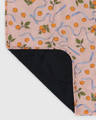 Die BAGGU Puffy Picknickdecke von Main Design hat einen pfirsichfarbenen Hintergrund mit einem orangefarbenen und grünen Fruchtmuster, Akzente aus blauen Bändern und eine Ecke, die gefaltet ist, sodass die schwarze Unterseite sichtbar ist. Aus recyceltem Nylon für Nachhaltigkeit gefertigt.