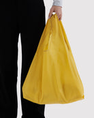 Eine Person trägt die BAGGU Standard-Tragetasche von BAGGU aus recyceltem Ripstop-Nylon in Gelb an den Henkeln, kombiniert mit einer schwarzen Hose und einem hellen Oberteil, vor einem schlichten, hellen Hintergrund.