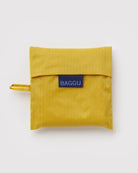 Eine zusammengefaltete BAGGU Standard-Tragetasche aus gelbem, recyceltem Ripstop-Nylon mit Klappenverschluss und blauem „BAGGU“-Label wird vor weißem Hintergrund präsentiert.