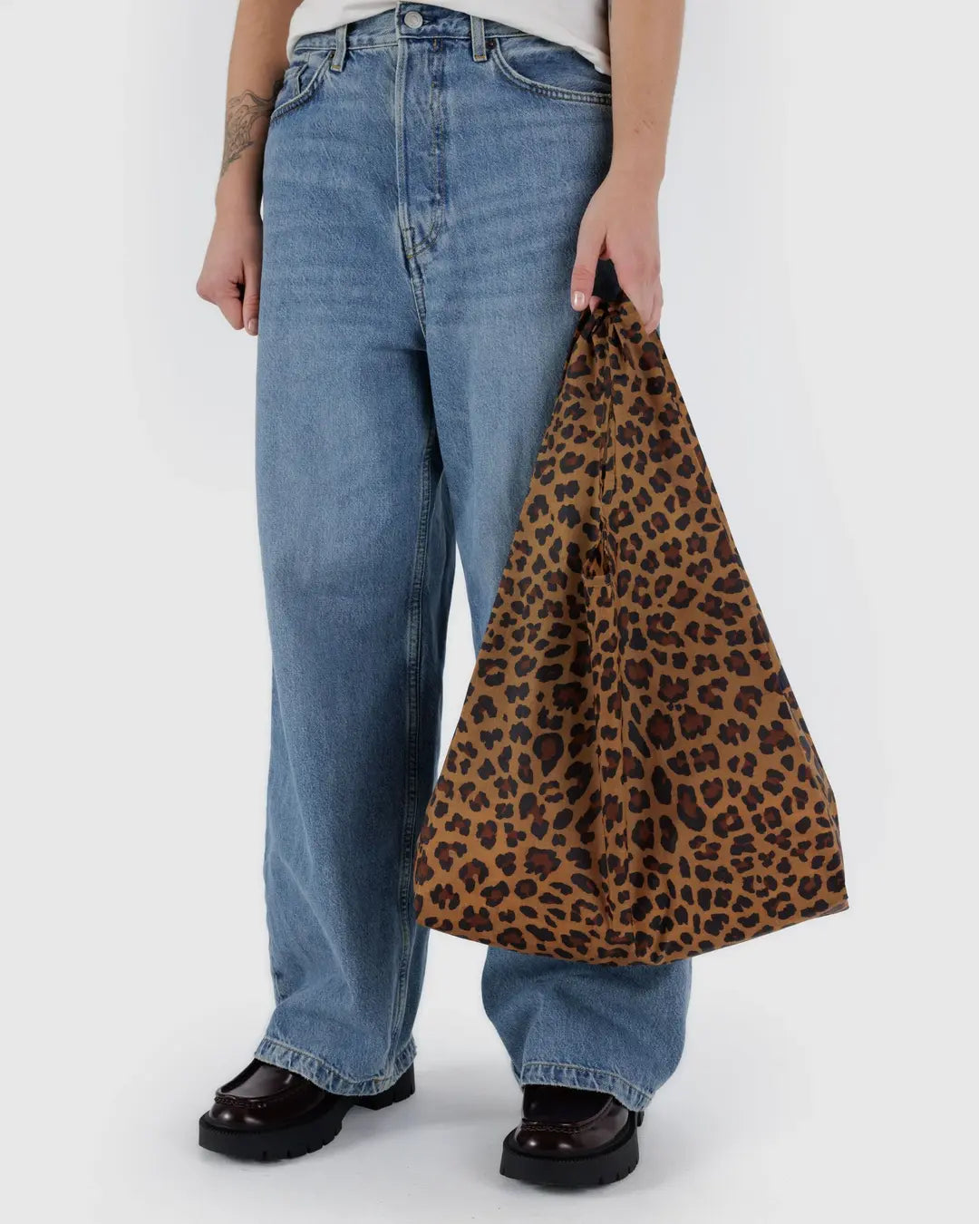 Eine Person in weiten Bluejeans hält eine BAGGU Standard Tragetasche von Main Design mit Leopardenmuster aus recyceltem Ripstop-Nylon. Sie trägt ein weißes Hemd und dunkelbraune Schuhe mit dicken Sohlen vor einem schlichten weißen Hintergrund.