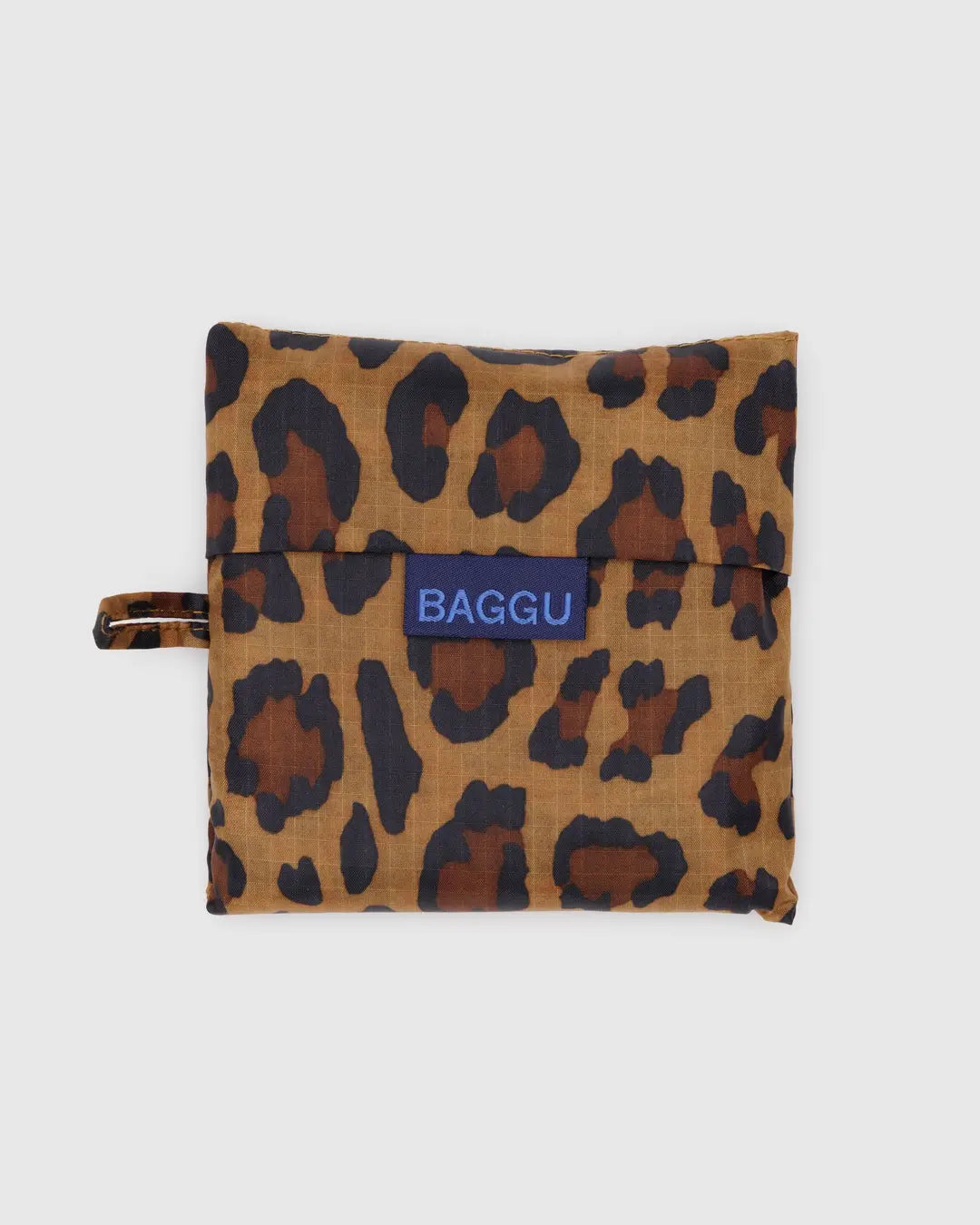 Eine gefaltete, wiederverwendbare BAGGU Standard-Tragetasche von Main Design zeigt ein Leopardenmuster auf hellgrauem Ripstop-Nylon mit einem Griff auf der linken Seite und einem blauen Etikett auf der Vorderseite mit der Aufschrift „Baggu“, was den umweltbewussten Einsatz recycelter Materialien durch die Marke unterstreicht.