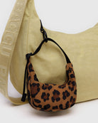 Ein kleiner BAGGU-Taschenanhänger in Form eines Halbmondes mit Leopardenmuster ist am Riemen einer größeren, hellgelben BAGGU-Tasche befestigt, die sich durch strukturierten Stoff und einen geprägten „BAGGU“-Schriftzug auf dem Riemen auszeichnet.