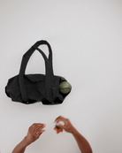 Eine BAGGU Everyday Cloud Bag von BAGGU ruht auf einer weißen Fläche und hält eine Melone. Zwei Hände - eine mit einer orangefarbenen Frucht - strecken sich nach dieser leichten Alltagstasche aus, die ein praktisches Laptop-Fach für die Dinge des täglichen Bedarfs enthält.