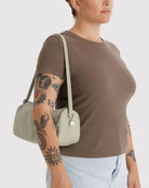 Eine Person mit Armtattoos trägt ein braunes T-Shirt und hellblaue Jeans und hat eine BAGGU Nylon Loaf Bag in Hellbeige mit silbernen Beschlägen dabei. Die Szene hat einen schlichten, hellen Hintergrund.