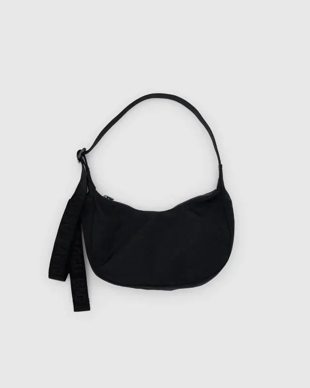 Die BAGGU Small Nylon Crescent Bag von Main Design ist eine kompakte schwarze Umhängetasche aus recyceltem Nylon in Halbmondform mit Reißverschluss, einzelnem Schultergurt und zwei dekorativen Quasten an den Seiten vor einem hellgrauen Hintergrund.