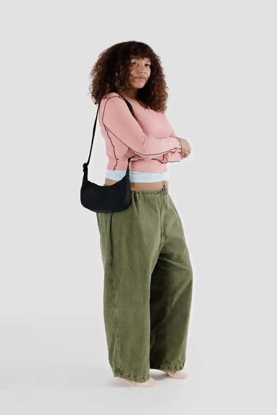 Eine Person mit lockigem Haar trägt ein rosa Langarmshirt und weite grüne Hosen und steht vor einem schlichten Hintergrund. Sie hat einen entspannten Gesichtsausdruck und trägt die BAGGU Small Nylon Crescent Bag von Main Design aus recyceltem Nylon.