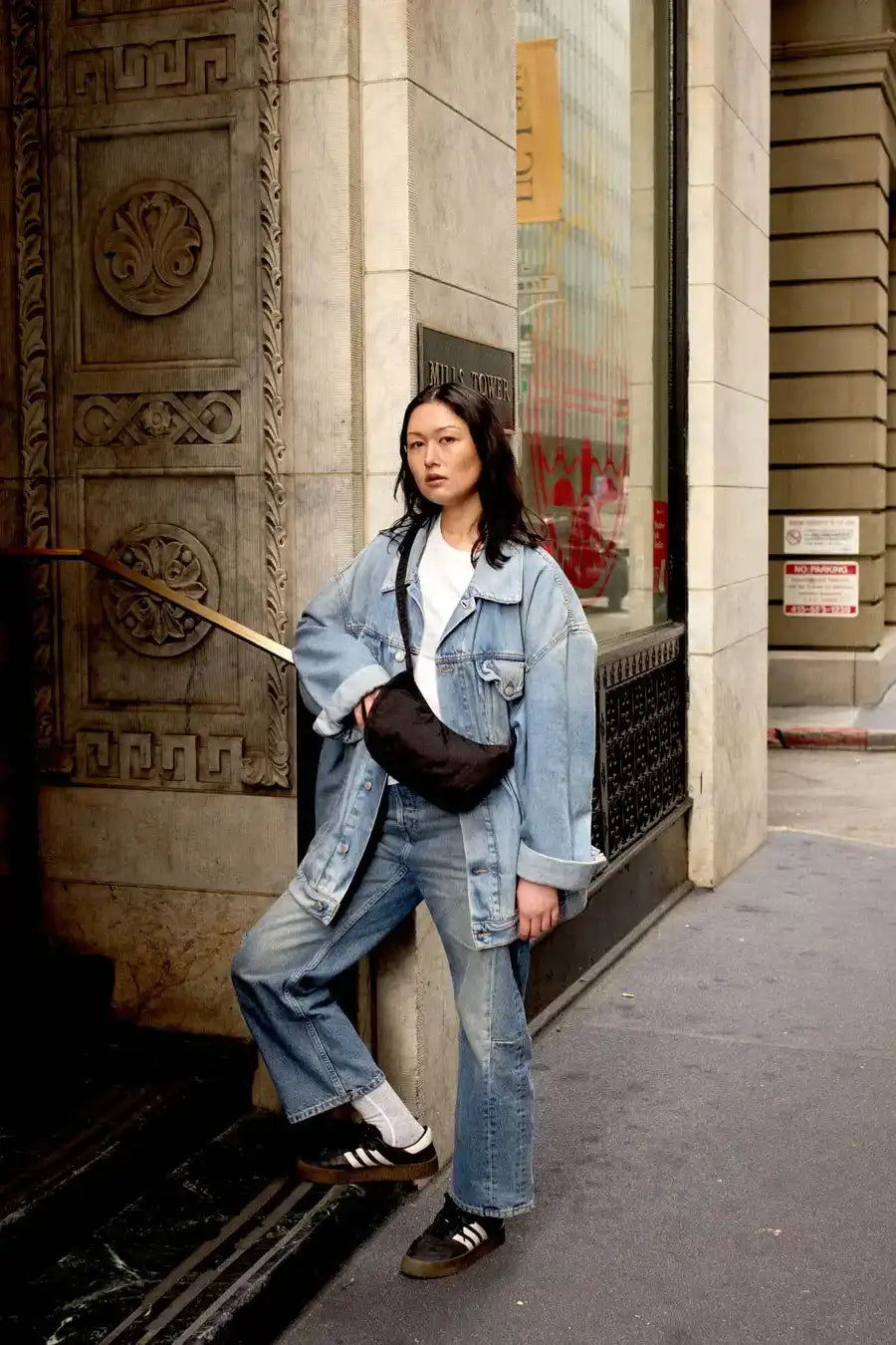 Eine Person in Jeansjacke, Jeans, weißem T-Shirt und schwarzen Adidas-Sneakern lehnt an einem Steingebäude. Sie hat schulterlanges Haar und trägt eine kompakte schwarze BAGGU Small Nylon Crescent Bag von Main Design. Die Szene zeigt urbane Straßenarchitektur.