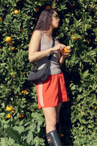 In einem grauen Tanktop, roten Shorts, schwarzen Stiefeln und einer Sonnenbrille schält eine Person neben einem üppigen Baum eine Orange. Über den Körper geschlungen trägt sie die BAGGU Small Nylon Crescent Bag von Main Design aus recyceltem Nylon.