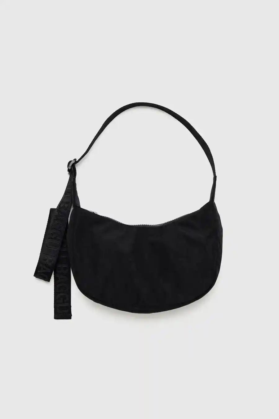 Die BAGGU Small Nylon Crescent Bag von Main Design ist eine schwarze Handtasche mit geschwungener Silhouette und einem einzelnen Riemen aus recyceltem Nylon. Sie verfügt über ein geprägtes, baumelndes Etikett, das vor einem schlichten weißen Hintergrund das umweltfreundliche Engagement der Marke unterstreicht.