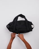 Die Hände einer Person öffnen eine schwarze BAGGU Small Cloud Carry-On Tasche von Main Design aus recyceltem Nylon auf weißem Grund. Die Tasche hat zwei Griffe und bietet Platz für Gegenstände, wobei auf jeder Seite eine Wasserflasche sichtbar ist.