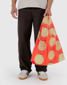Eine Person mit dunkler Hose und weißen Sneakern hält die BAGGU Standard Tragetasche von Main Design aus recyceltem Ripstop-Nylon mit gelbem Apfeldesign vor hellem Hintergrund.