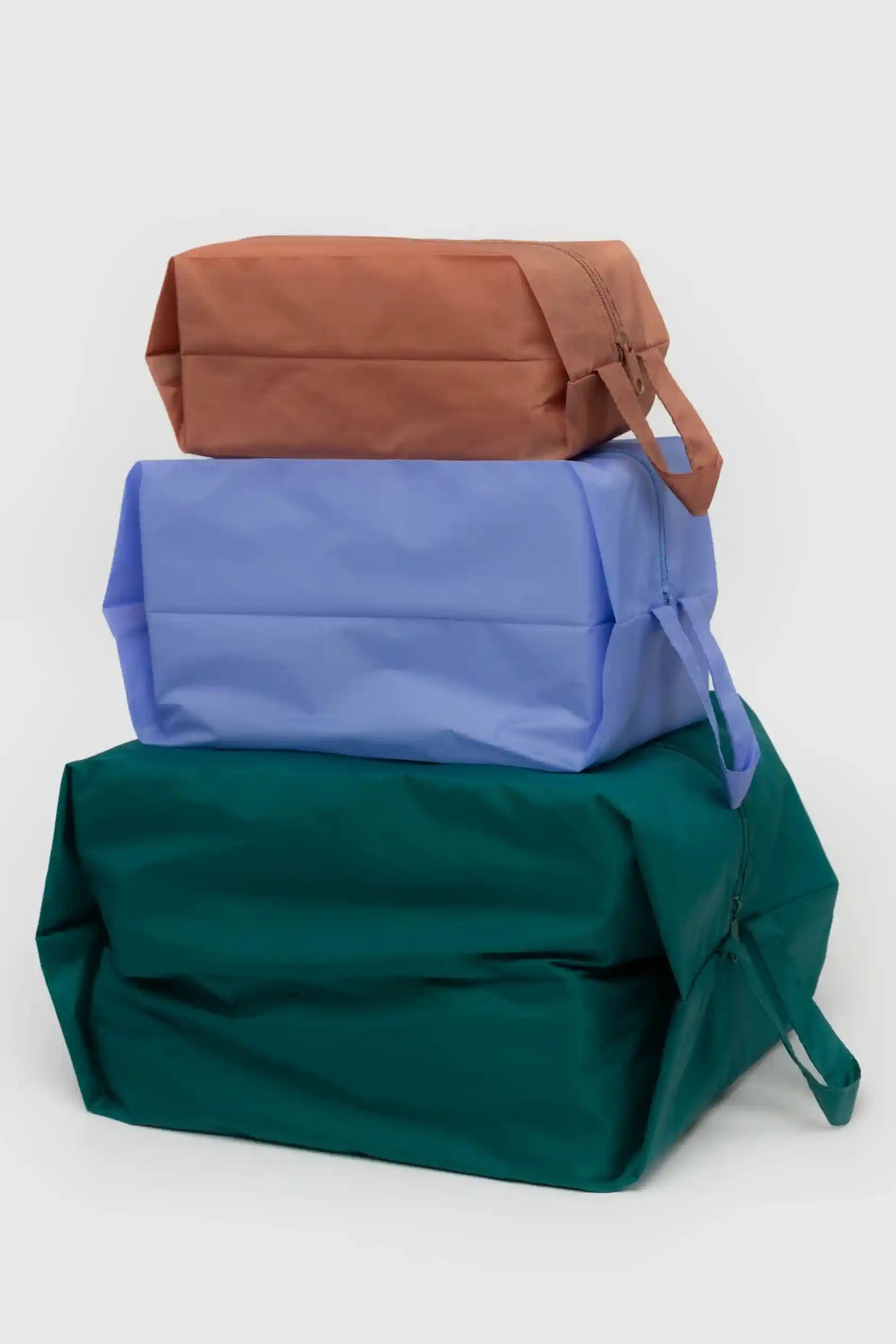 Ein Stapel aus drei rechteckigen BAGGU 3D Zip Set-Beuteln von Main Design aus recyceltem Ripstop-Nylon, übereinander angeordnet. Die obere Tasche ist braun und hat einen Griff, die mittlere Tasche ist hellblau und die untere Tasche ist dunkelgrün. Alle drei Taschen zeichnen sich durch ein einfaches, schmuckloses Design auf einem schlichten hellen Hintergrund aus.