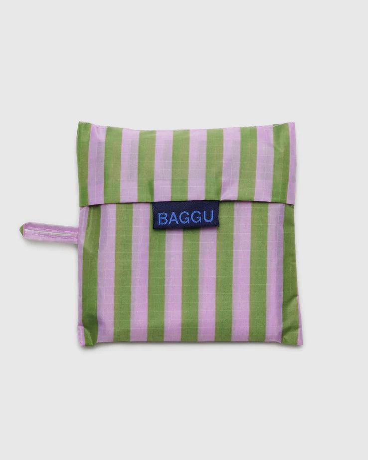 Die BAGGU Standard Tragetasche von Main Design ist in einem rechteckigen Beutel mit Klappe untergebracht. Der Beutel ist mit vertikalen grünen und rosa Streifen versehen und hat eine kleine Schlaufe auf der linken Seite. Die Klappe besteht aus recyceltem Ripstop-Nylon und trägt ein blaues Schild mit dem Text „BAGGU“ in schwarzen Buchstaben, das ein umweltbewusstes Unternehmen präsentiert.