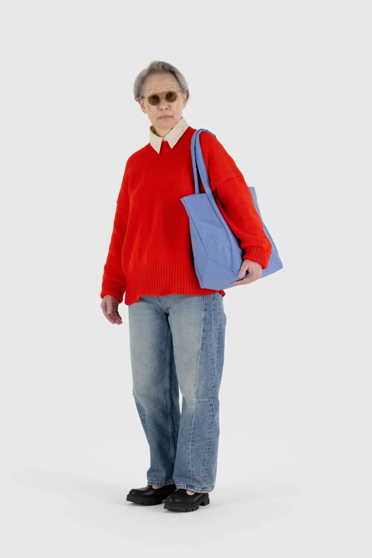 Eine Person mit kurzen grauen Haaren steht vor einem schlichten weißen Hintergrund. Sie trägt eine Sonnenbrille, einen leuchtend roten Pullover über einem weißen Hemd, blaue Jeans, schwarze Schuhe und trägt die Main Design BAGGU Cloud Bag aus recyceltem Nylon auf der linken Schulter.