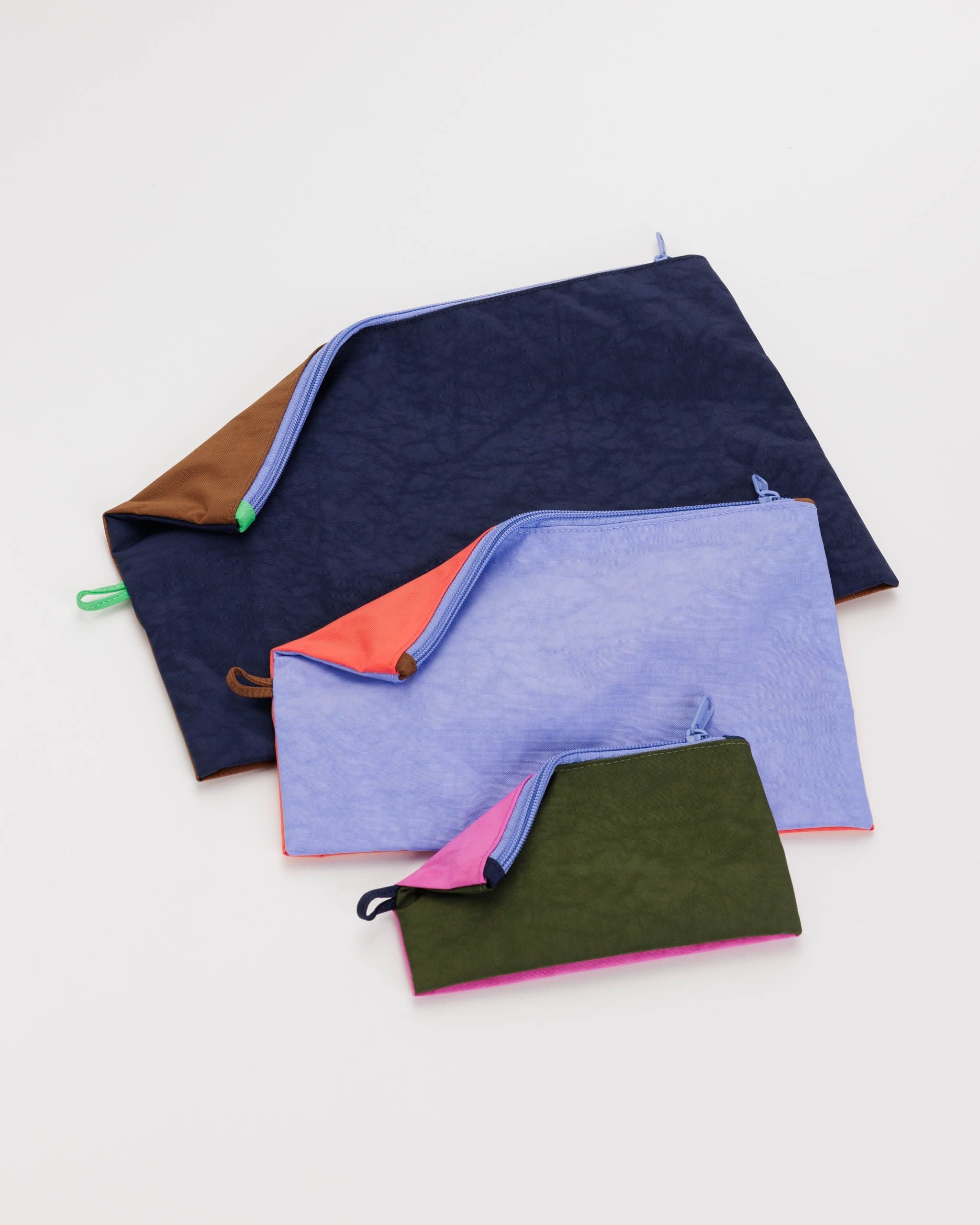 Das Main Design BAGGU Go Pouch Set mit drei Reißverschlusstaschen in unterschiedlichen Größen ist ordentlich auf einem weißen Hintergrund angeordnet. Die größte Tasche aus recyceltem Nylon ist dunkelblau mit einem braunen und grünen Reißverschluss, die mittlere Tasche ist hellviolett mit einem korallenfarbenen und rosa Reißverschluss und die kleinste Tasche ist grün mit einem schwarzen und rosa Reißverschluss.