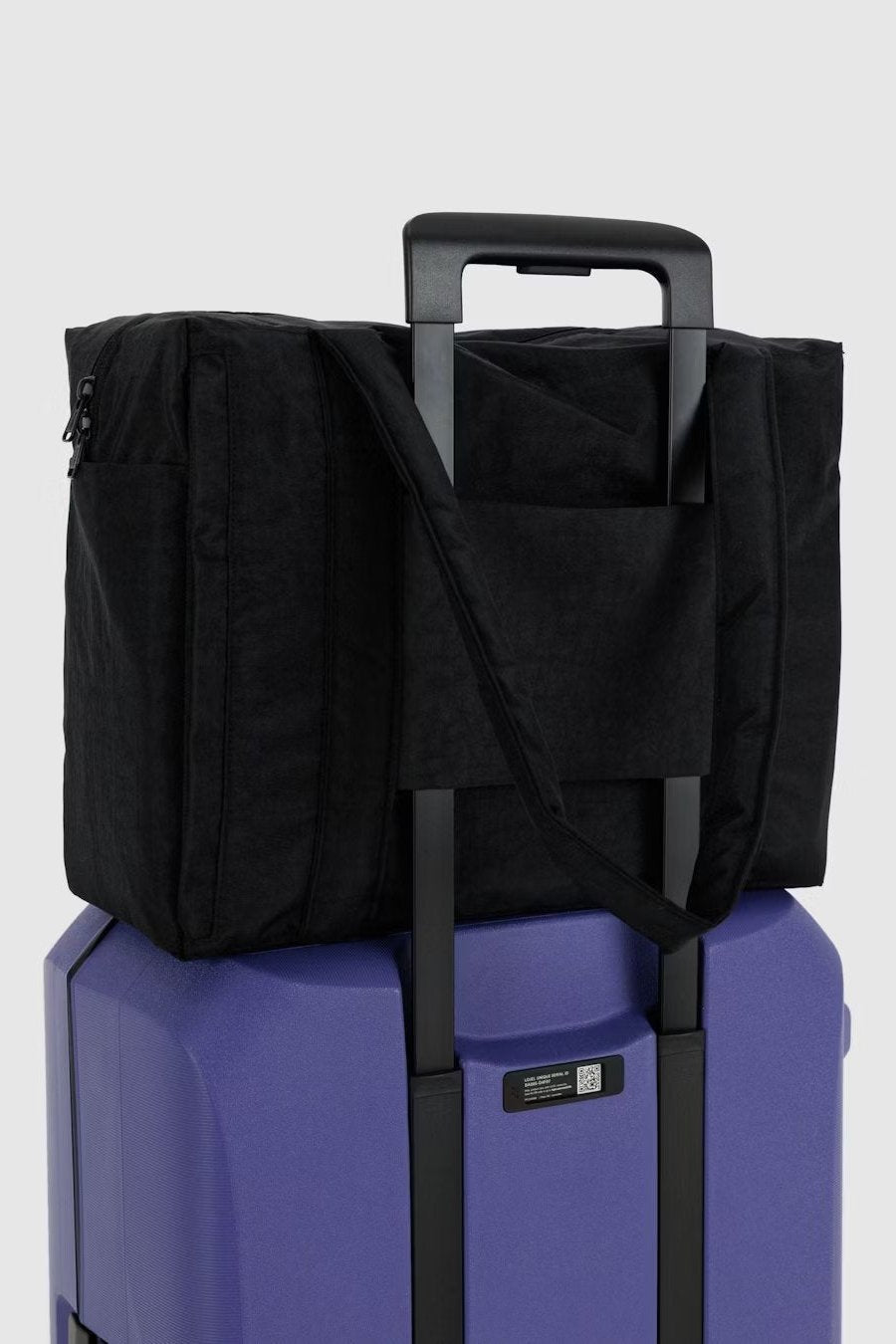 Eine Main Design BAGGU Small Cloud Carry-On Tasche in Schwarz sitzt sicher auf einem lila Koffer. Die Tragegurte der Tragetasche sind über den verlängerten Koffergriff geschlungen. Der schlichte, helle Hintergrund betont sowohl das Gepäck als auch die BAGGU Tasche.