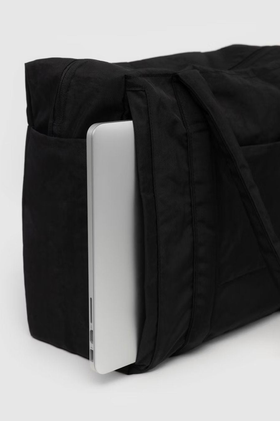 Nahaufnahme einer schwarzen BAGGU Small Cloud Carry-On Tasche von Main Design mit offener Seitentasche, in der ein silberner Laptop teilweise eingelegt ist. Der Hintergrund ist schlicht und hell und hebt die Tasche und den Laptop hervor.