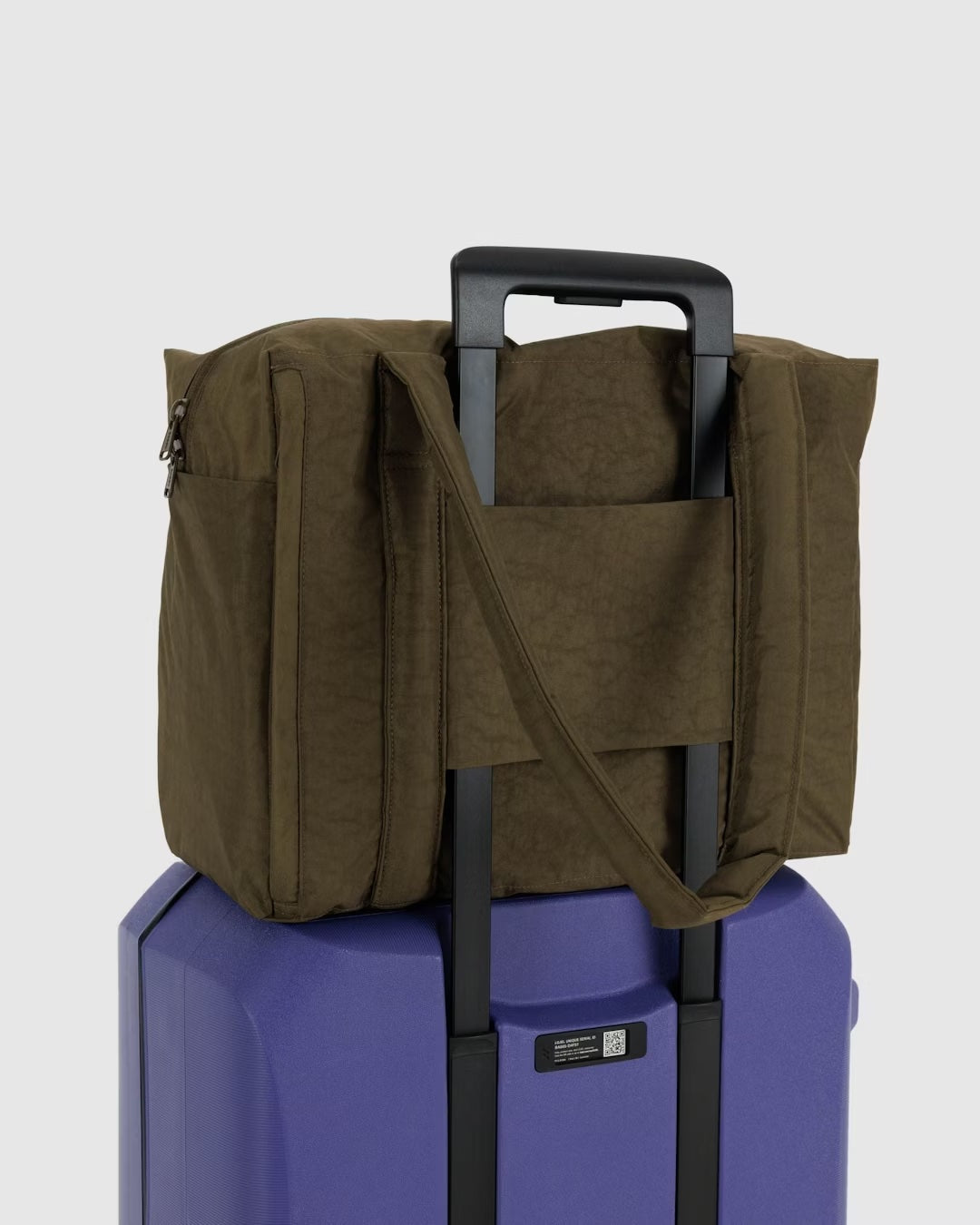 An einem violetten Hartschalenkoffer mit verlängertem Griff ist eine Main Design BAGGU Small Cloud Carry-On Tasche in Olivgrün sicher befestigt, indem man sie über den Griff schiebt. Die Reisetasche verfügt über einen Reißverschluss und Tragegurte. Der Hintergrund ist schlicht weiß.